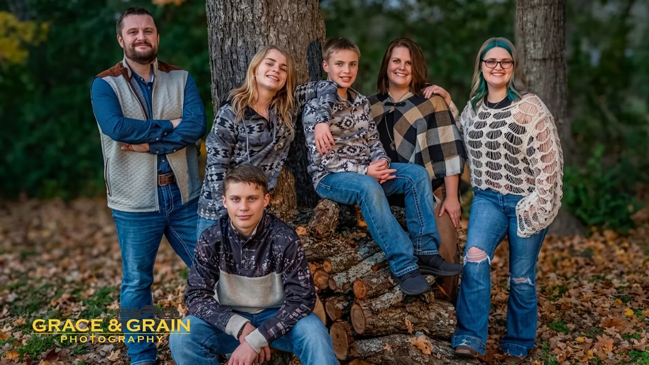 A family of six gathered by a woodpile in fall leaves